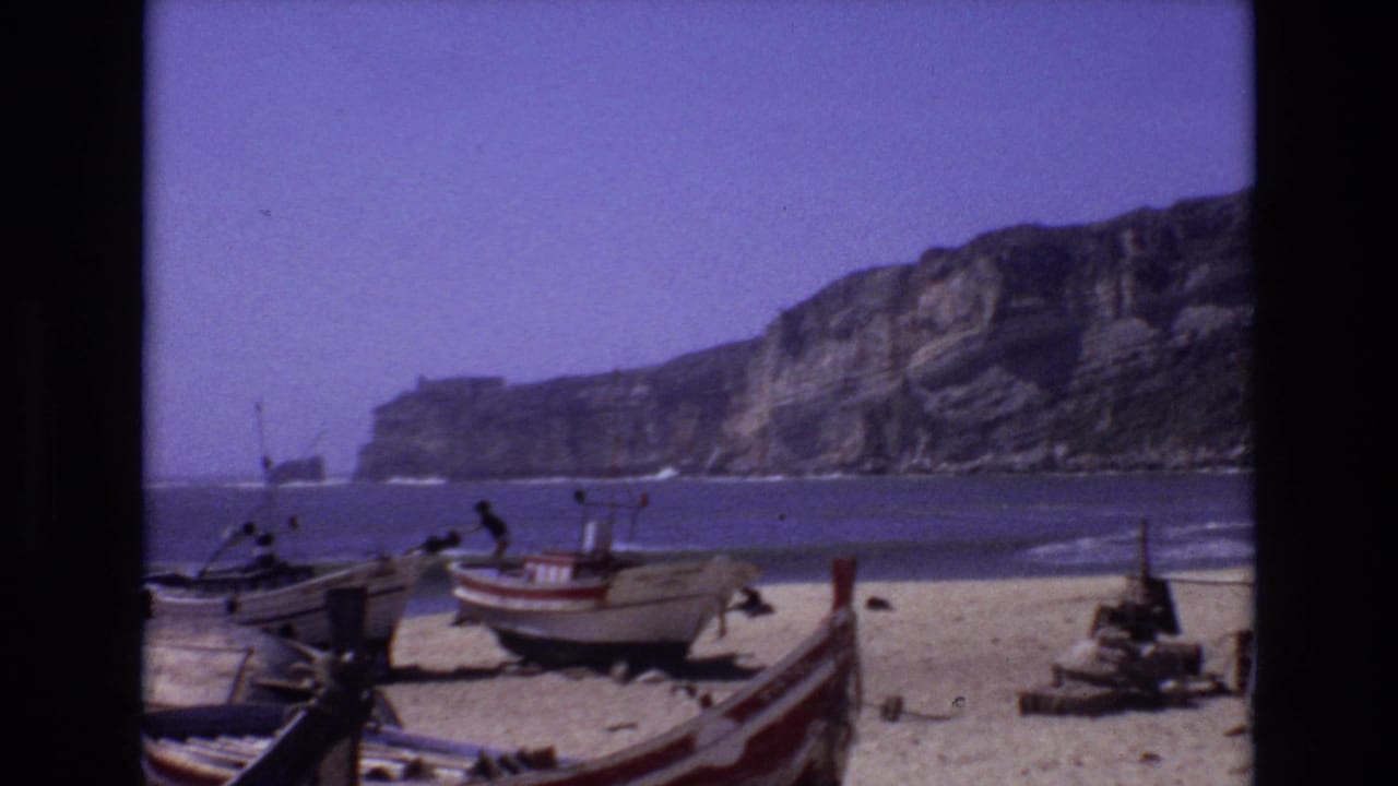 1972 Portugal 8mm frame showing small beach boats on sand with waves, distant people, and high coastal cliffs in the background.