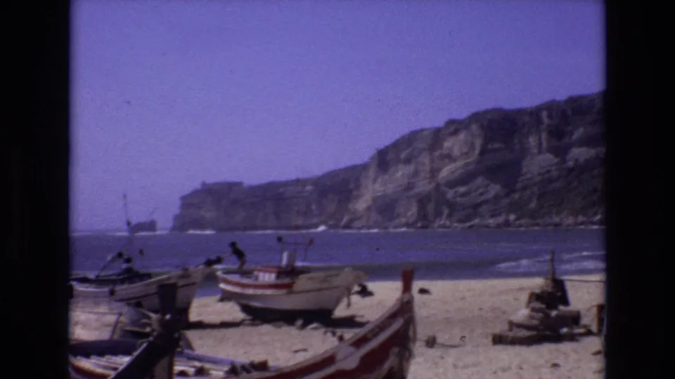 1972 Portugal 8mm frame showing small beach boats on sand with waves, distant people, and high coastal cliffs in the background.