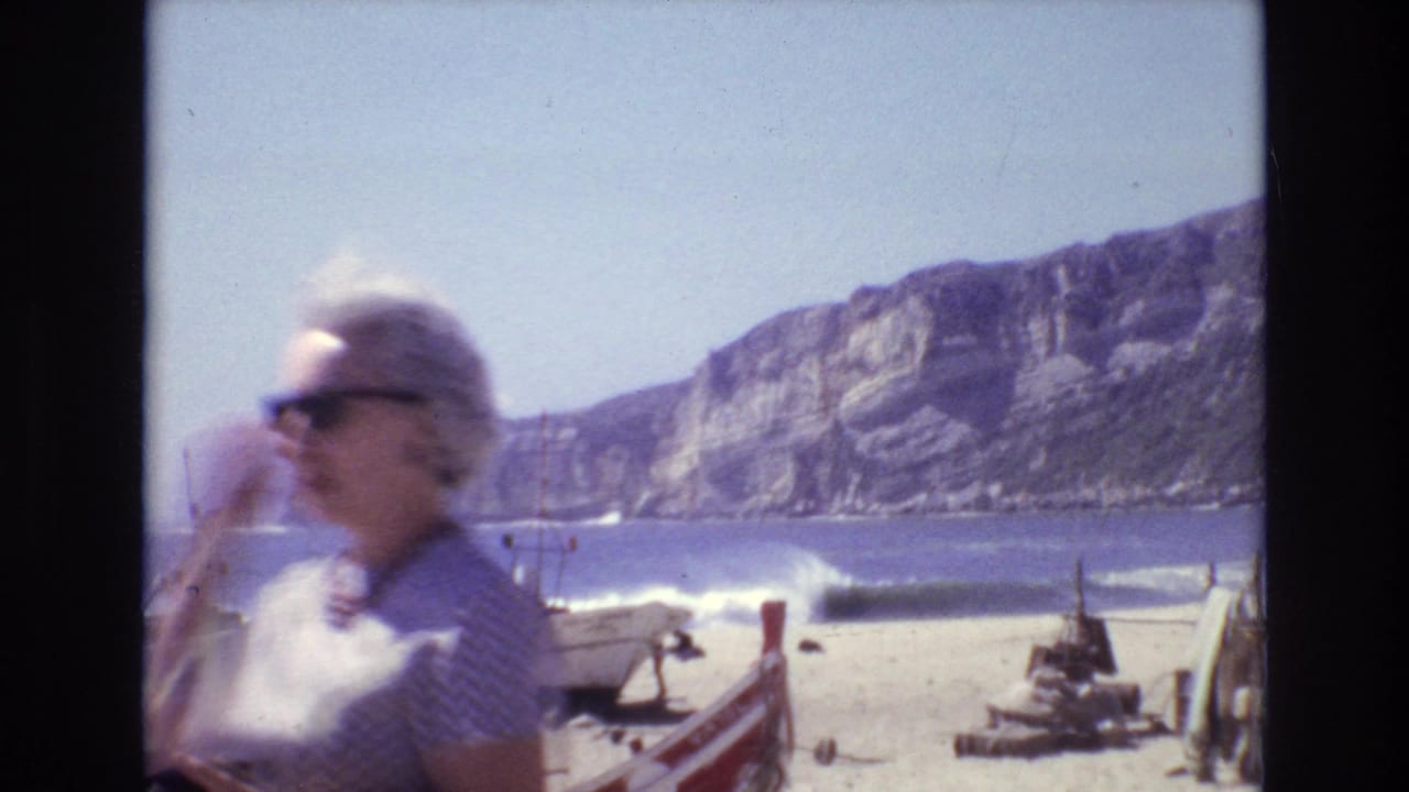 A blonde woman in sunglasses passes in front of boats on a beach by cliffs.