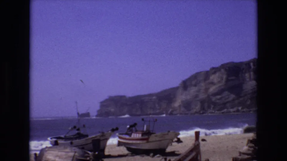 1972 Portugal 8mm frame showing small boats on a sandy shore, waves breaking nearby, and rocky coastal cliffs across the bay.