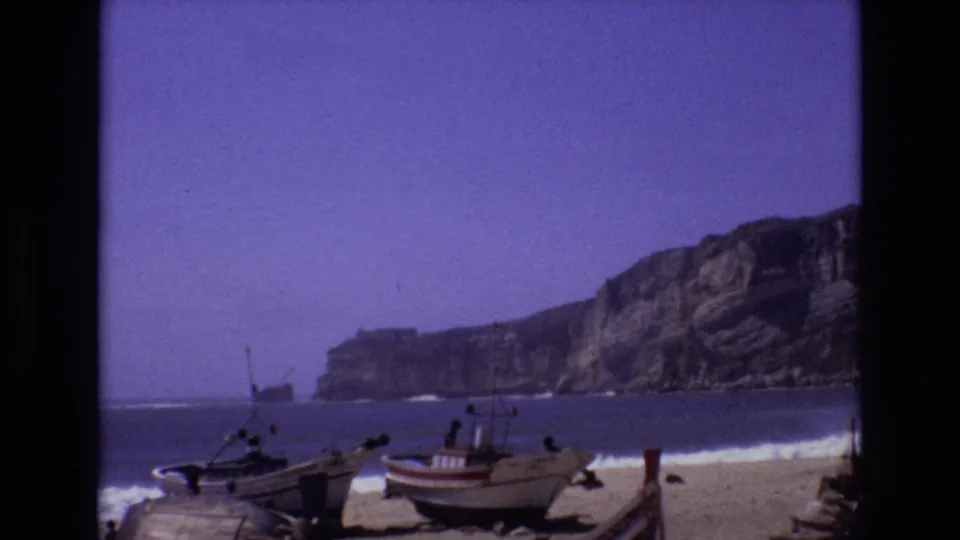 1972 Portugal 8mm frame of beach boats near the surf, with distant people, rocky cliffs, and a purple-tinted sky in home-movie color.