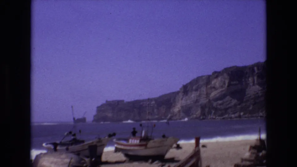 1972 Portugal 8mm frame showing small fishing boats on sand beside breaking surf, with cliffs rising along the coast under a hazy sky.