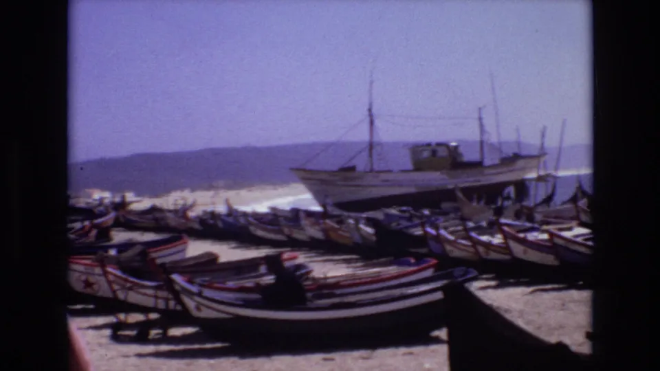 1972 Portugal coastal frame with many small boats lined on sand and a larger fishing vessel set back near the surf, with distant hills across the water.