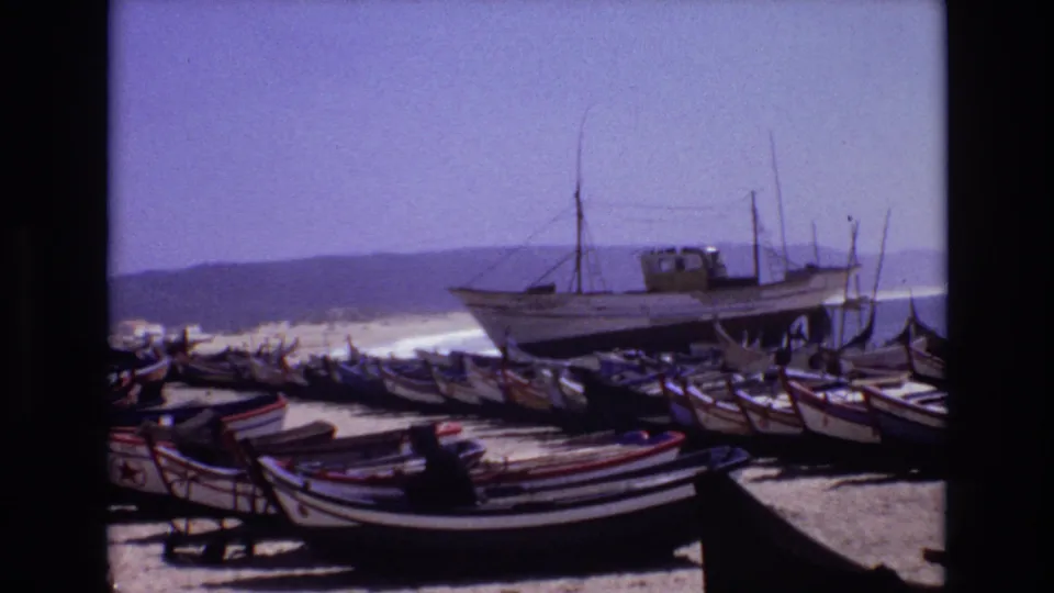 1972 Portugal 8mm still of a crowded fishing beach, with striped small boats in the foreground, a larger vessel behind them, and waves breaking near the shore.