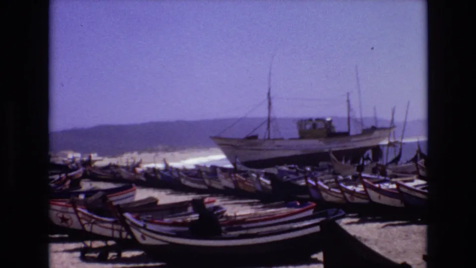 1972 Portugal frame showing a group of beached fishing boats and a larger vessel against surf and hazy hills, with the soft grain and color shift of 8mm film.