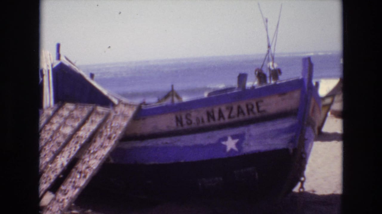1972 Portugal 8mm frame showing a weathered blue wooden fishing boat marked NS DA NAZARE on sand beside the surf.