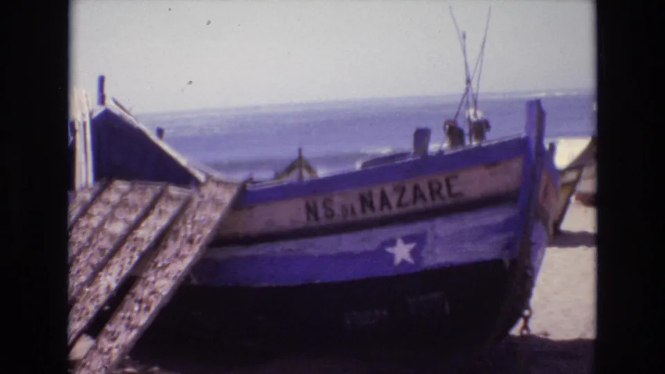 1972 Portugal 8mm frame showing a weathered blue wooden fishing boat marked NS DA NAZARE on sand beside the surf.