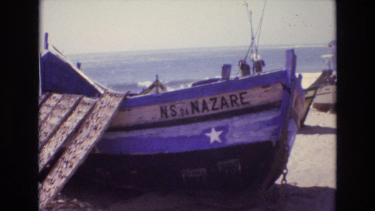 A blue wooden fishing boat marked NS DA NAZARE sits angled on a Portuguese beach, with surf and small distant figures visible behind it.