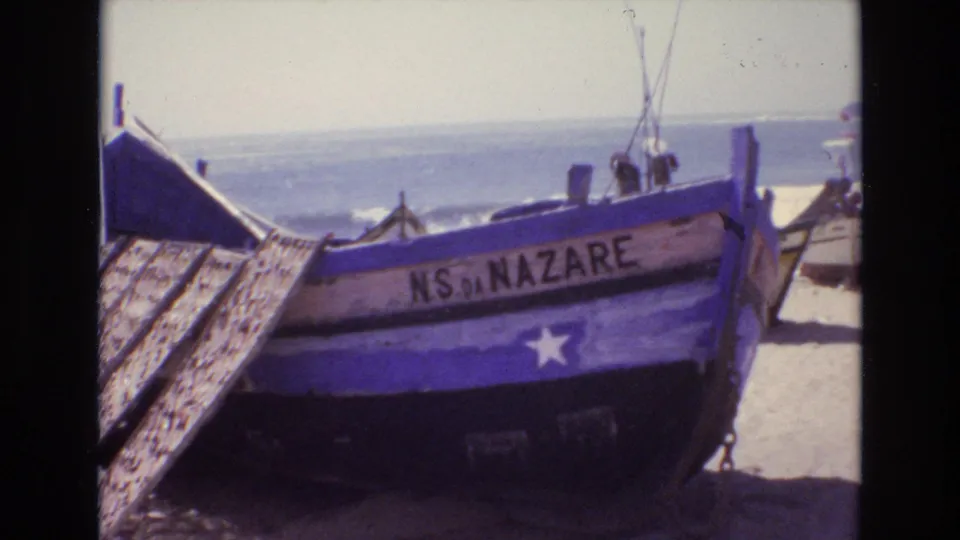 A blue wooden fishing boat marked NS DA NAZARE sits angled on a Portuguese beach, with surf and small distant figures visible behind it.