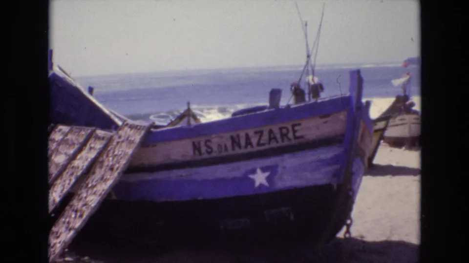 Home-movie frame from Portugal in 1972 showing the blue NS DA NAZARE boat, wooden beach planks, sand, and Atlantic waves beyond.