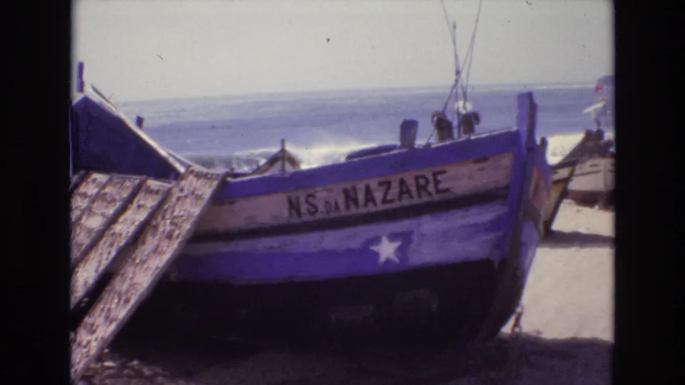 The final frame holds on the weathered blue NS DA NAZARE fishing boat on a Portuguese beach, with waves, sky, and nearby boat details visible.
