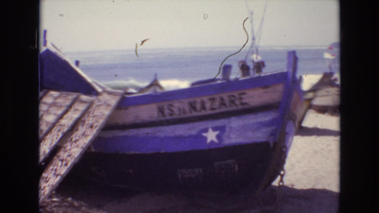 A blue and white wooden fishing boat labeled “NS DA NAZARE” sits on a sandy Portuguese beach in 1972, with the ocean visible beyond it.