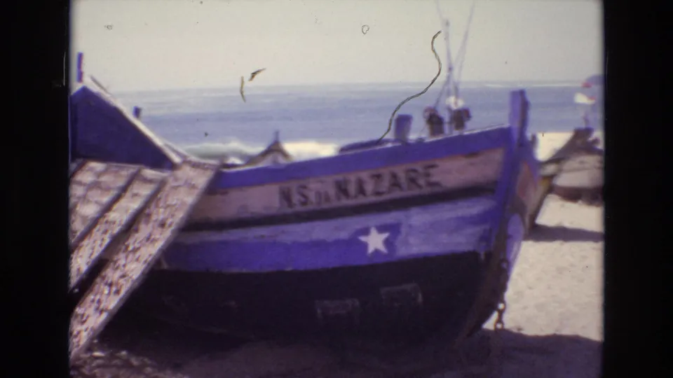 A blue and white wooden fishing boat labeled “NS DA NAZARE” sits on a sandy Portuguese beach in 1972, with the ocean visible beyond it.