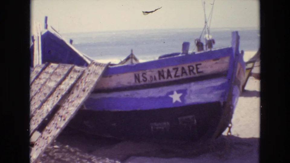 The beached wooden boat labeled “NS DA NAZARE” is framed closer, with weathered planks at left and surf in the background on a sunny Portuguese shore.