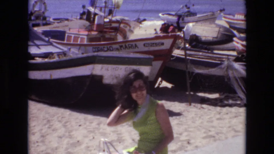 A woman in a green sleeveless dress and sunglasses poses on a sandy Portugal beach in 1972, with several beached fishing boats and painted registration markings behind her.