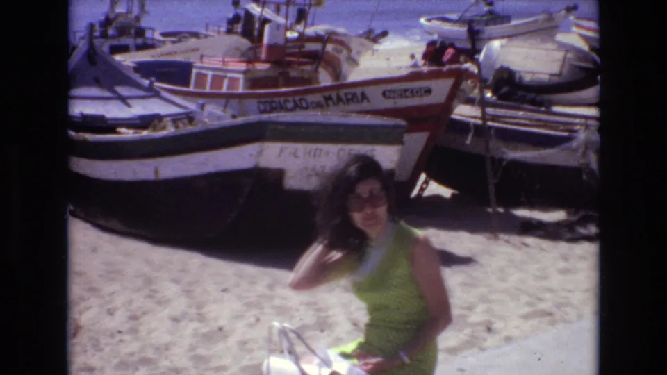 The woman remains in the foreground on the sandy shore, partially turned toward the camera, while clustered fishing boats fill the background in this 1972 Portugal home-movie frame.