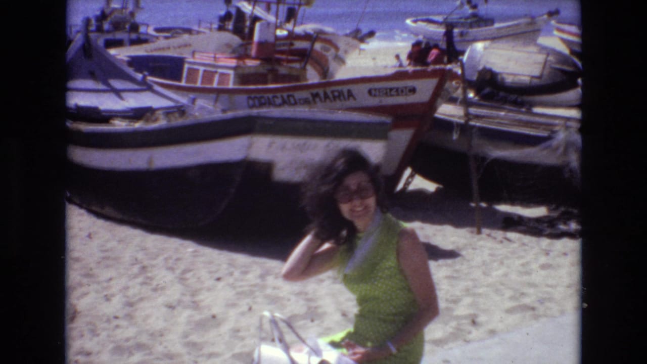 Woman in a green dress and sunglasses poses on a sandy beach in front of boats.