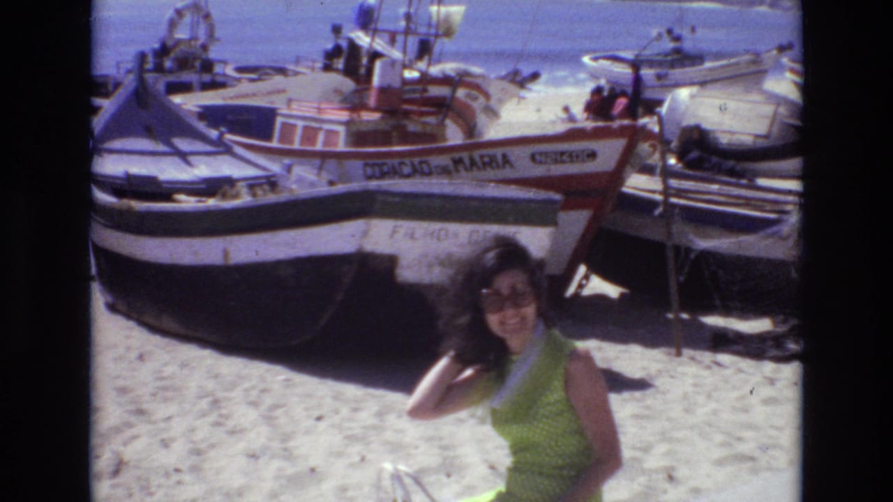 Woman smiles toward the camera beside fishing boats on a bright sandy beach.