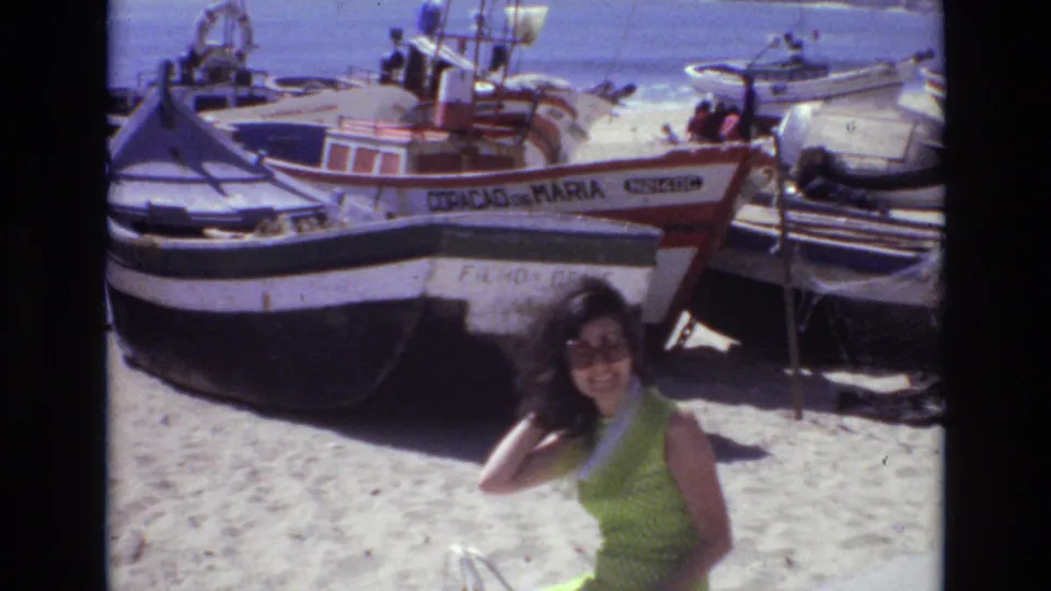A smiling brunette woman in sunglasses stands on a Portuguese beach in 1972, with beached boats and visible hull lettering behind her.