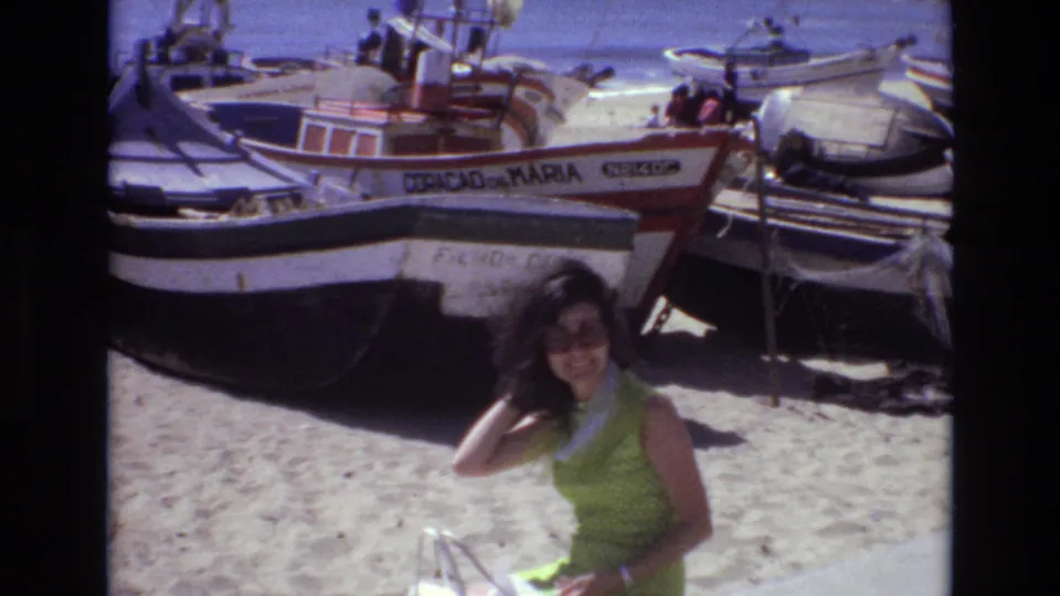 The woman adjusts her hair while posing on a sunny Portuguese beach in 1972, framed by several small boats resting on the sand.