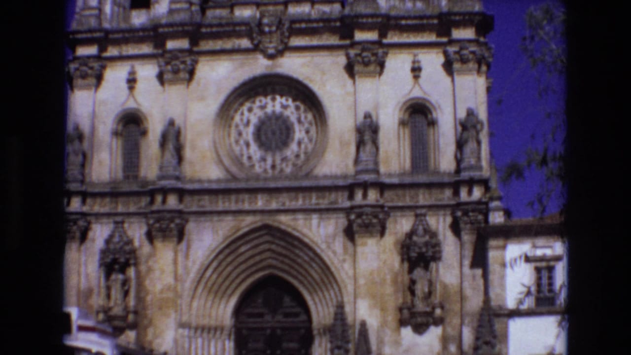 1972 Portugal 8mm frame showing the lower facade of an ornate historic church with a circular rose window, pointed arched entrance, carved stone figures, and blue sky.