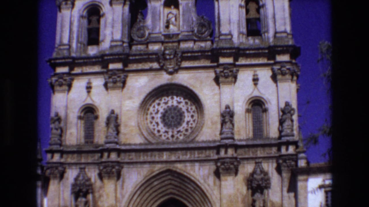 1972 Portugal 8mm frame showing an ornate church front with a rose window, upper niches, carved figures, and partial bell tower openings against a deep blue sky.