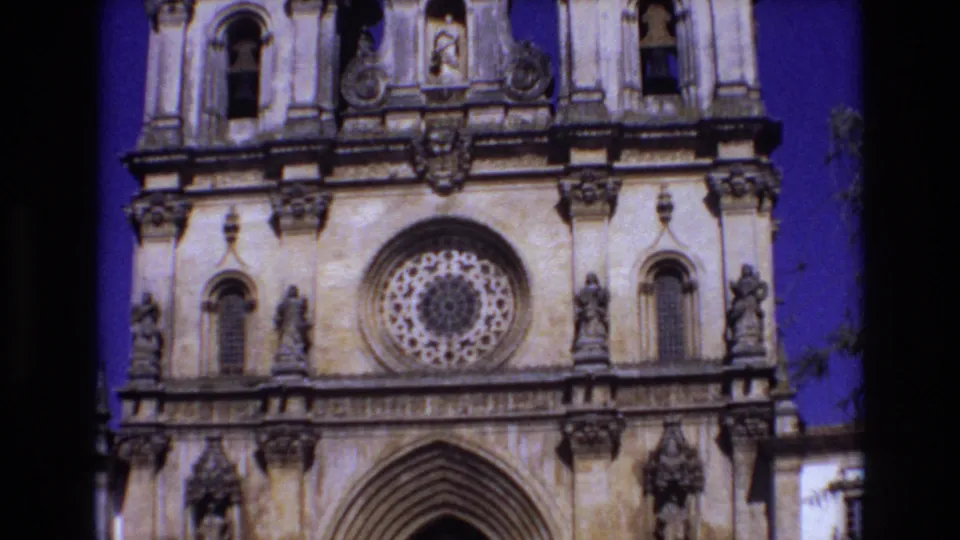 1972 Portugal 8mm frame showing an ornate church front with a rose window, upper niches, carved figures, and partial bell tower openings against a deep blue sky.