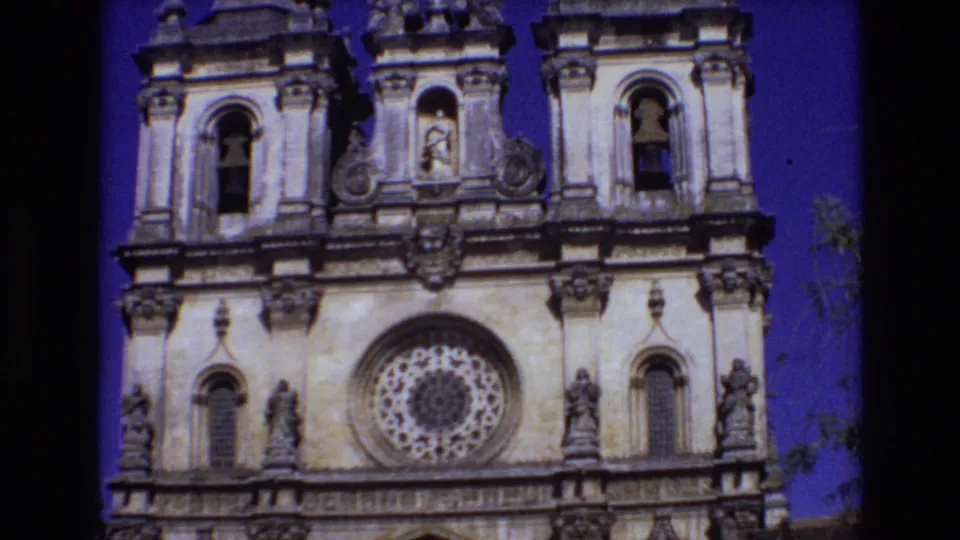 1972 Portugal 8mm frame of a historic church exterior, centered on the bell towers, sculpted stonework, rose window, and statues under a vivid blue-purple sky.