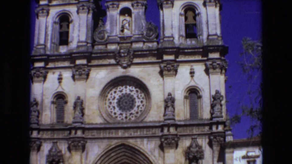 1972 Portugal 8mm frame with the camera tilted lower on the ornate church front, revealing the rose window, statues, arched doorway, bell towers, and a small edge of nearby foliage or building.