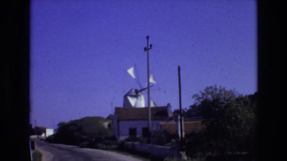 1972 Portugal 8mm frame showing a white windmill on low ground beside a road, with utility poles, trees, and a deep blue sky.