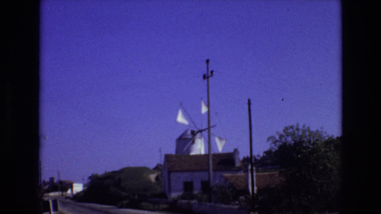 Portugal 1972 home-movie frame showing a white roadside windmill with sails, low buildings, utility poles, shrubs, and a deep blue-purple sky.