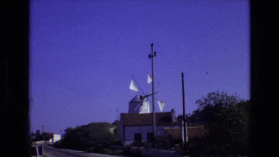 Portugal 1972 home-movie frame showing a white roadside windmill with sails, low buildings, utility poles, shrubs, and a deep blue-purple sky.