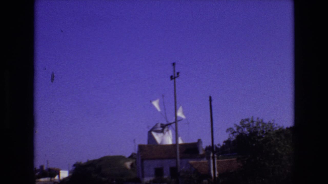 Portugal 1972 frame with the white windmill shifted slightly in view, its sails visible above a low structure, utility poles, roadside greenery, and open sky.