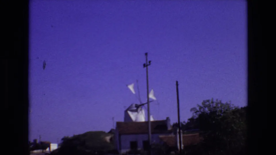 Portugal 1972 frame with the white windmill shifted slightly in view, its sails visible above a low structure, utility poles, roadside greenery, and open sky.