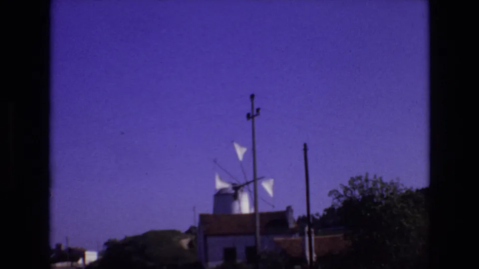 Portugal 1972 home-movie still showing the windmill partly behind roadside buildings, with white sails, utility poles, a shrub mass, and a saturated blue-purple sky.