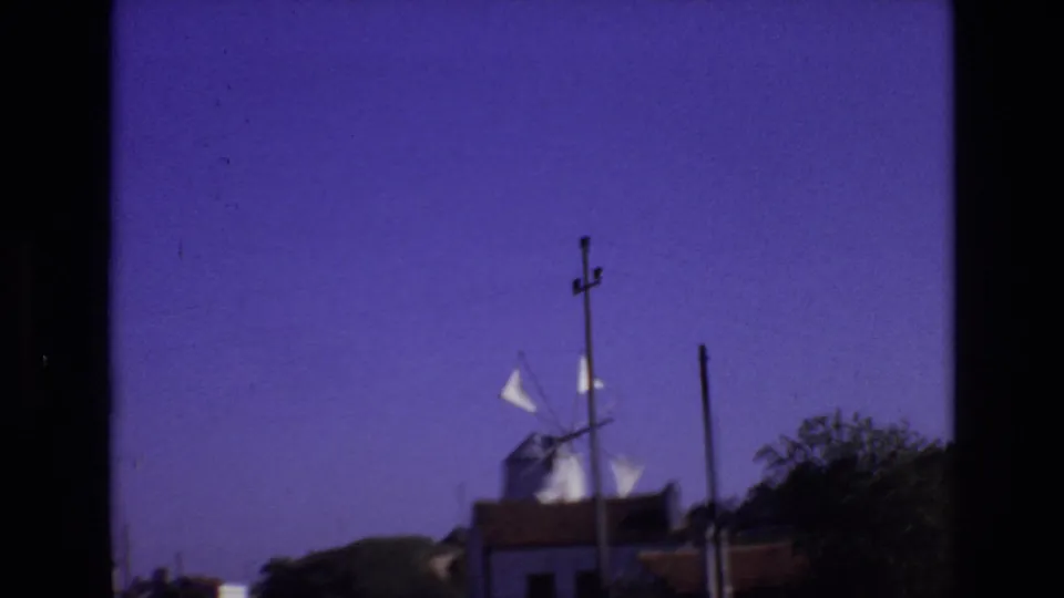 Portugal 1972 frame showing a distant white windmill with sails near low roadside buildings, utility poles, dark border edges, vegetation, and a clear blue-purple sky.