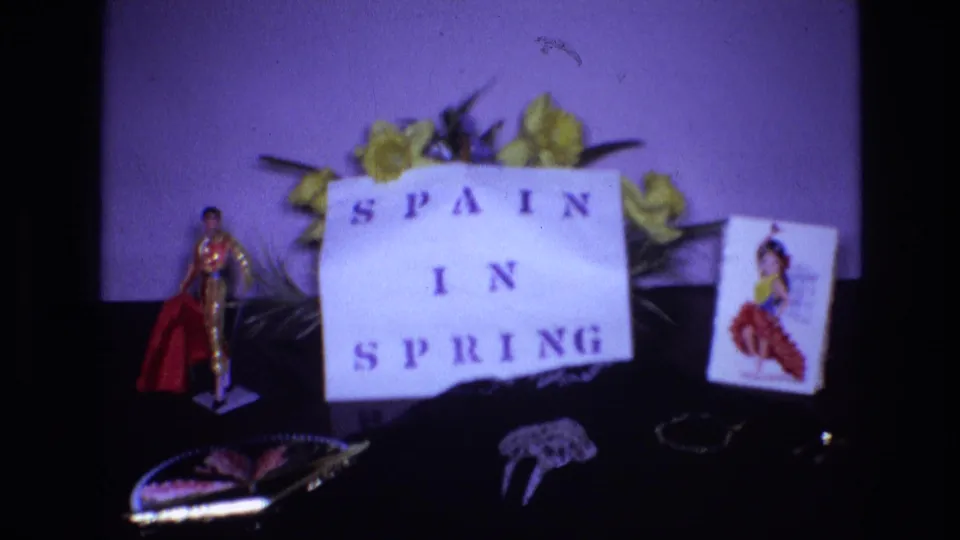 Spanish-themed 1972 home-movie still life with a handmade SPAIN IN SPRING sign surrounded by yellow flowers, a bullfighter figurine, a folding fan, jewelry or hair pieces, and a card showing a dancer.