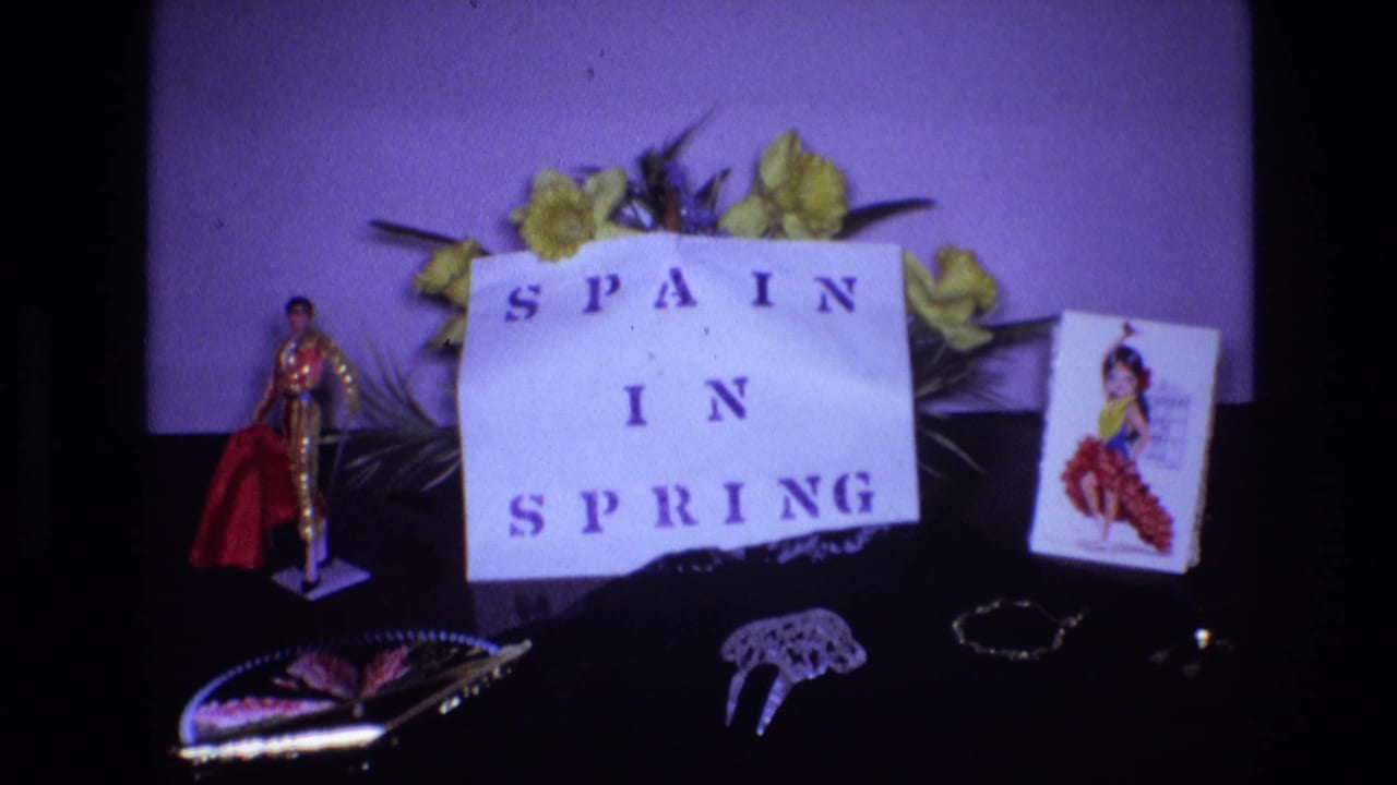 Paper sign reading "SPAIN IN SPRING" surrounded by flowers, figurine, postcard, fan, and jewelry-like objects.