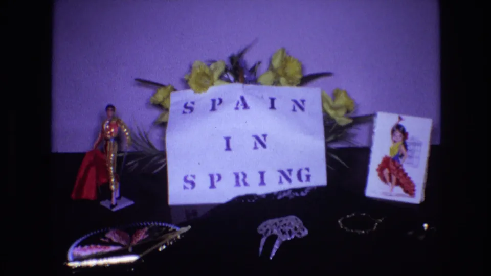 1972 Spain-themed home-movie still life showing the handmade sign, yellow flowers, small bullfighter figure, flamenco dancer card, fan, lace object, and bracelet under a purple film cast.