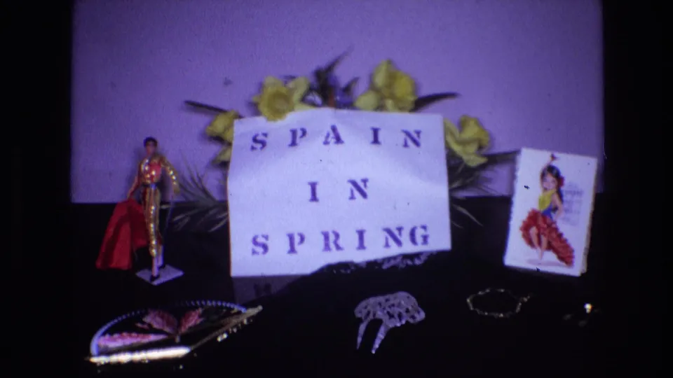 Soft 8mm frame from 1972 showing a closer Spanish-themed tabletop arrangement with the paper "SPAIN IN SPRING" sign, flowers, souvenir figure, dancer card, and decorative objects.