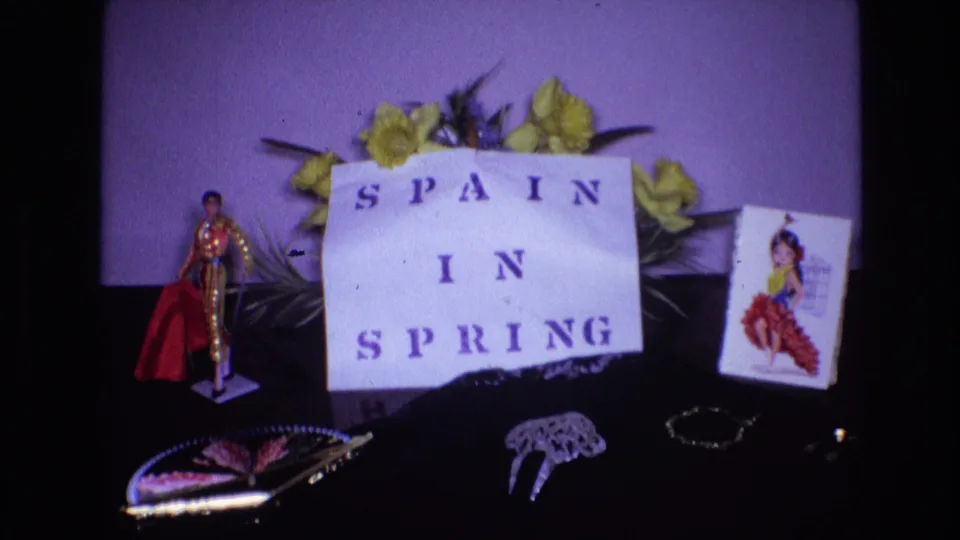 Final 1972 8mm frame of the Spanish souvenir display, with the "SPAIN IN SPRING" sign centered between yellow flowers, a bullfighter figurine, a flamenco dancer card, and jewelry-like decorations.