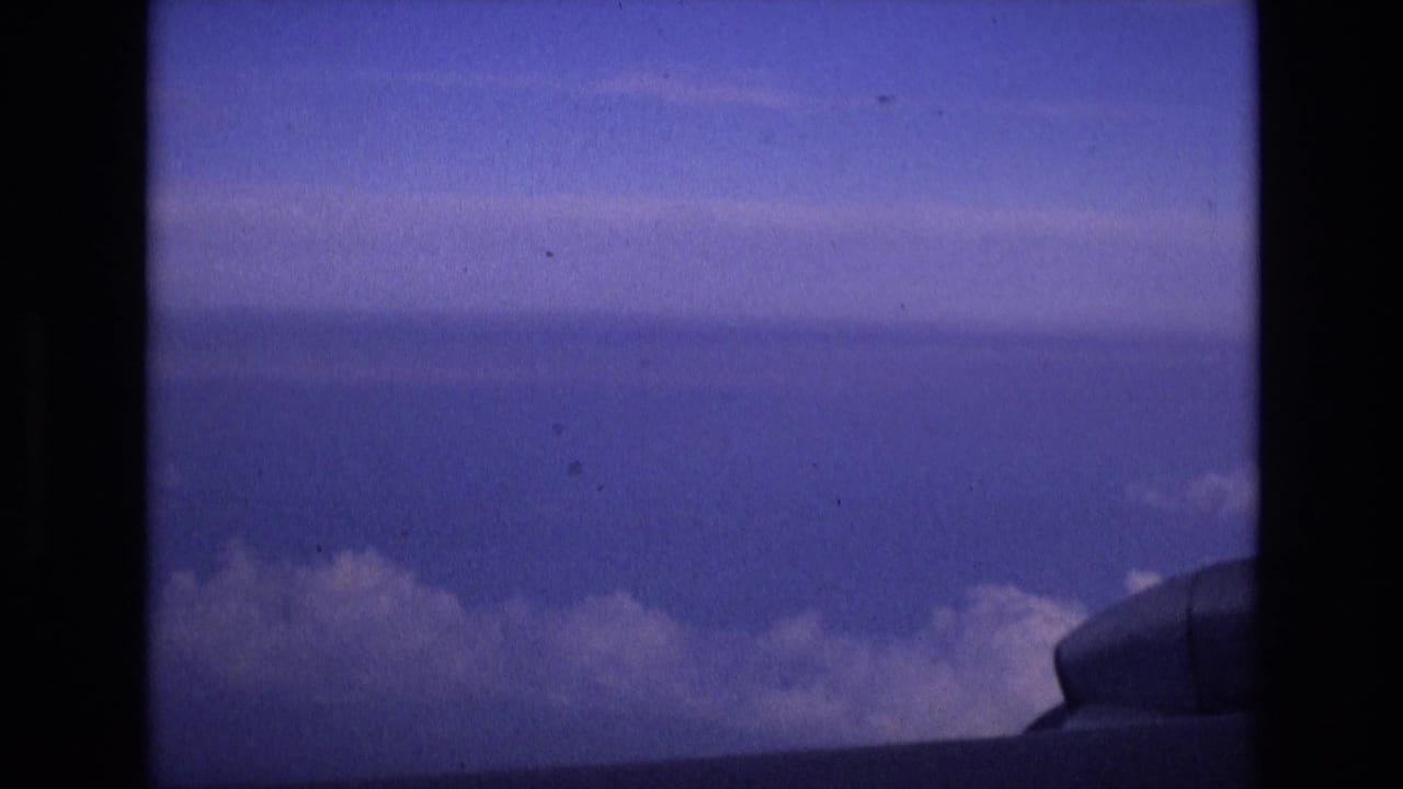 A 1972 Spain-associated 8mm frame shows blue sky, a horizontal cloud layer, and part of the aircraft structure near the lower right, viewed from a passenger-window perspective.