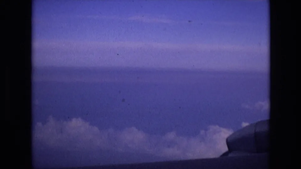 A 1972 Spain-associated 8mm frame shows blue sky, a horizontal cloud layer, and part of the aircraft structure near the lower right, viewed from a passenger-window perspective.