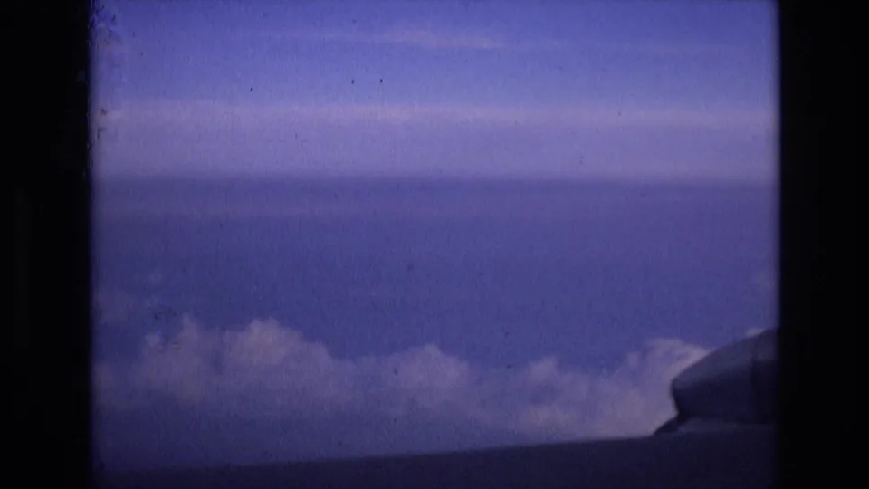The frame shows a steady passenger-window view over clouds, with blue sky above and a small portion of aircraft structure visible at the lower right in 1972 Spain-associated home-movie footage.