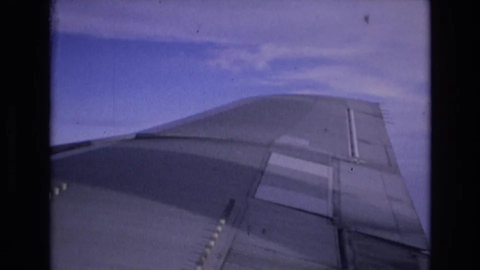 1972 Spain 8mm frame showing an airplane wing from a passenger-window view, with blue sky and light clouds above the wing.