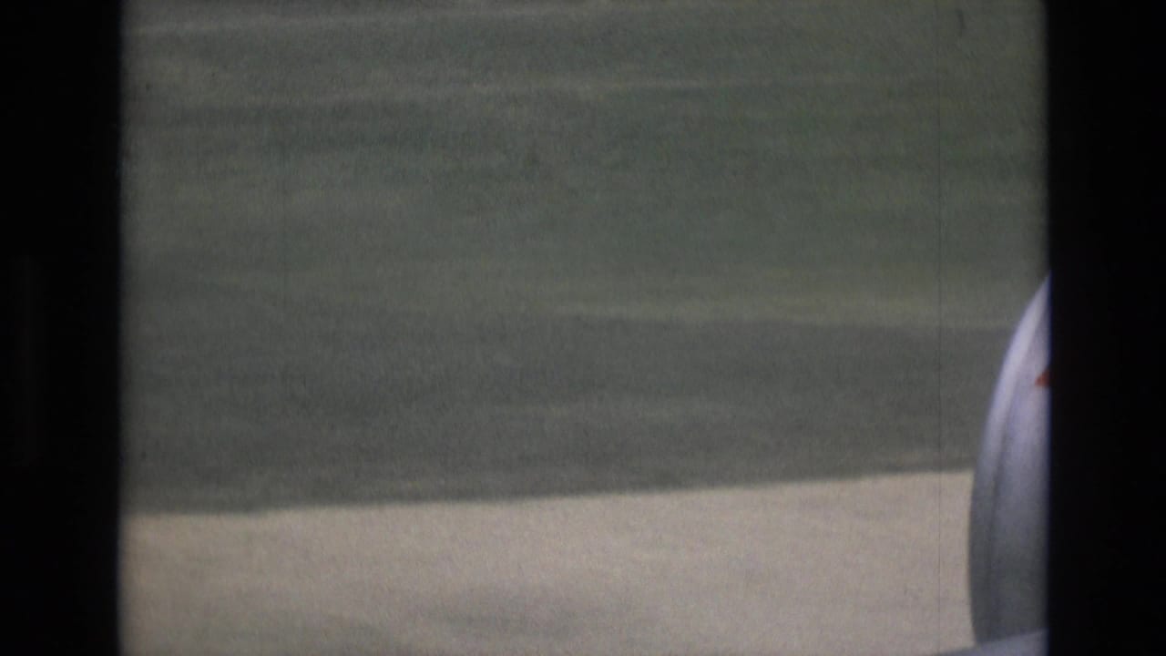 1972 Spain 8mm passenger-window frame showing blurred ground, a pale runway or taxiway edge, and part of the aircraft engine at the side of the view.