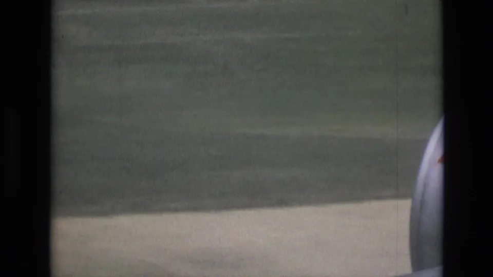 1972 Spain 8mm passenger-window frame showing blurred ground, a pale runway or taxiway edge, and part of the aircraft engine at the side of the view.