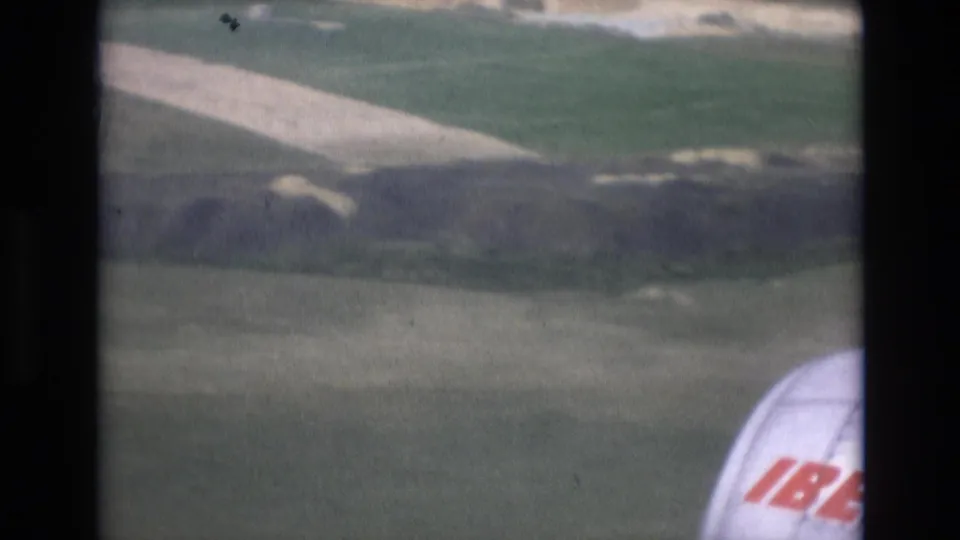 Aircraft descends over airport-edge fields in Spain, with a pale runway or service strip below and partial Iberia lettering visible on the aircraft at right.