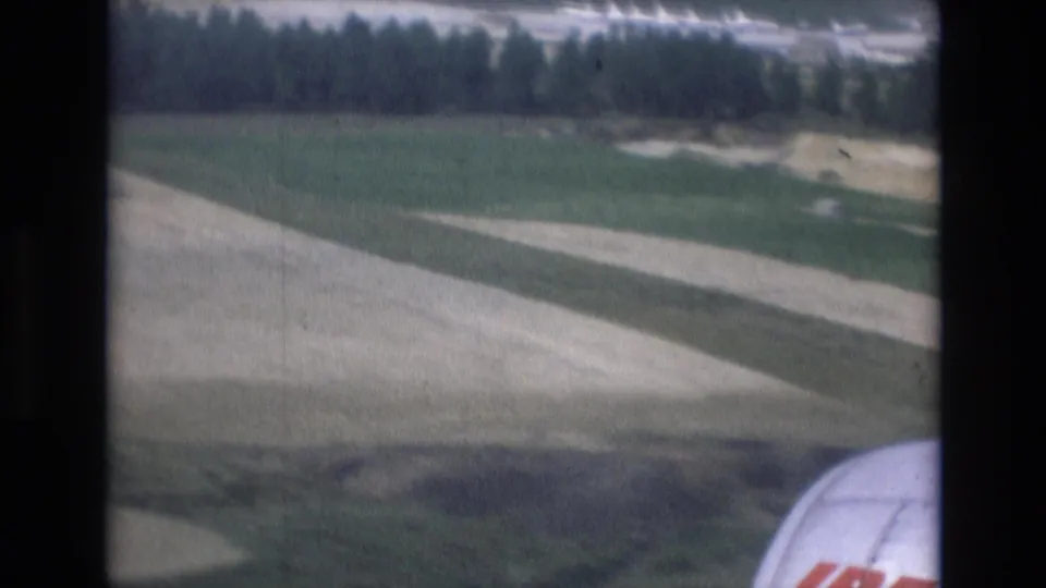 Low passenger-window view during a 1972 Spain landing approach, showing a pale runway or taxiway strip, fields, distant trees, and part of the aircraft at lower right.