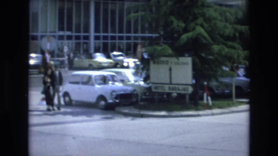 Street-level 1972 Spain view near Hotel Barajas with parked cars, pedestrians, trees, luggage, and a modern glass-fronted building.